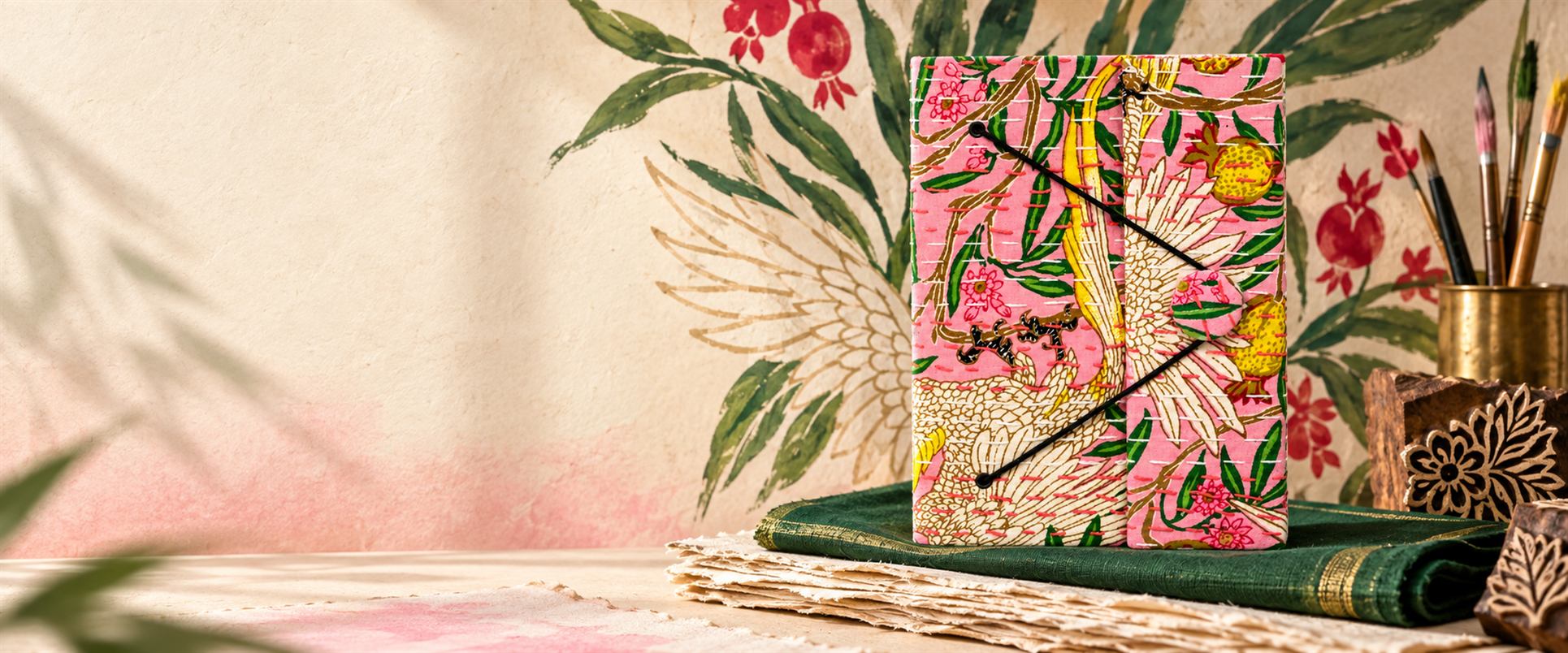 A pink block-printed fabric journal with a black cord closure, standing on green cloth and handmade paper beside a brass pot of paintbrushes and carved wooden printing blocks